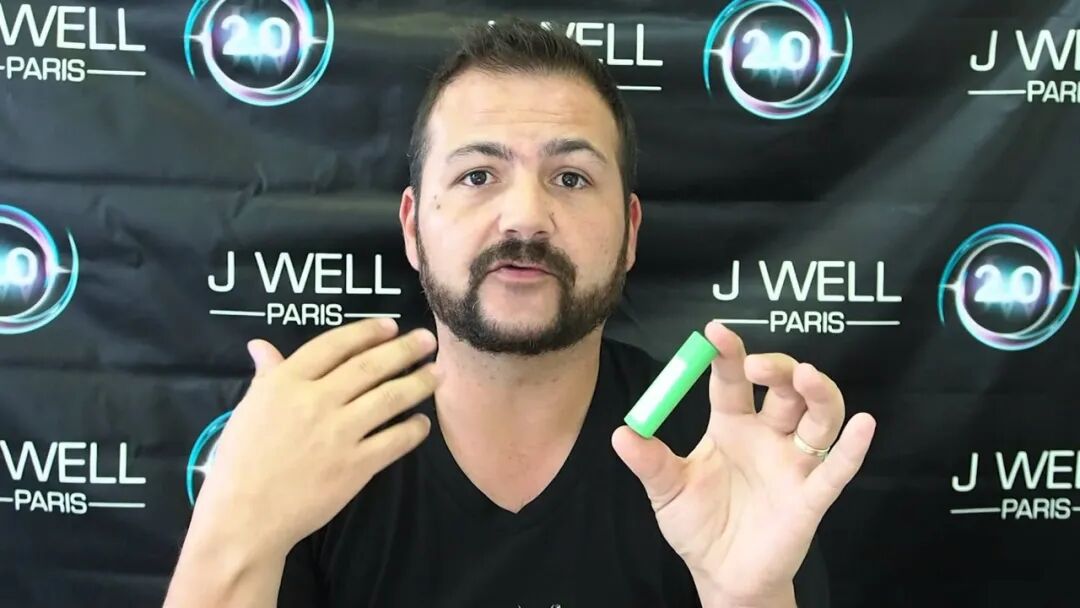 一名男子站在印有 J WELL PARIS 的背景前，手持一支綠色裝置。